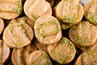 Circular cookies with a light brown color are stacked closely together. Each cookie has distinct horizontal ridges on the surface and is sprinkled with green bits, possibly crushed pistachios or herbs.