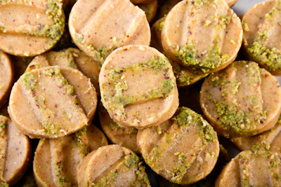 Circular cookies with a light brown color are stacked closely together. Each cookie has distinct horizontal ridges on the surface and is sprinkled with green bits, possibly crushed pistachios or herbs.