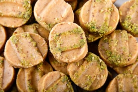 Circular cookies with a light brown color are stacked closely together. Each cookie has distinct horizontal ridges on the surface and is sprinkled with green bits, possibly crushed pistachios or herbs.