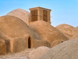 A series of traditional mud-brick domes are clustered together under a clear blue sky. The central focus is a box-like windcatcher structure with vertical wooden slats, rising above the domes, likely used for natural ventilation. The earthy tones of the domes contrast subtly against the sky, suggesting a harmonious blend of architecture with the natural environment.