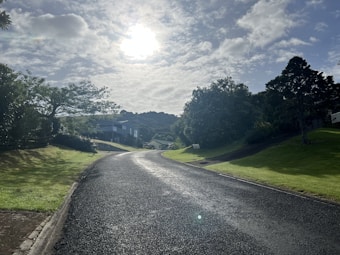 A sunlit suburban street curves gently upward, flanked by lush green lawns and trees. The sky is dotted with light clouds, allowing beams of sunlight to cast a serene glow over the landscape. Houses are partially visible in the background, nestled among the vegetation.