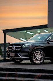 A black Mercedes-Benz GLS 350d is parked in front of a modern building during sunset. The sky features warm orange and yellow tones, providing a striking contrast to the sleek, dark vehicle. Architectural elements in the background include glass and metal structures, indicative of an urban setting. The car&rsquo;s LED headlights are illuminated, adding a touch of sophistication to the scene.