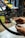 Hands are adjusting a bicycle disc brake with a tool, focusing on the intricate components of the brake system. The background includes a cluttered workshop environment with various tools and equipment visible.