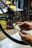 Hands are adjusting a bicycle disc brake with a tool, focusing on the intricate components of the brake system. The background includes a cluttered workshop environment with various tools and equipment visible.