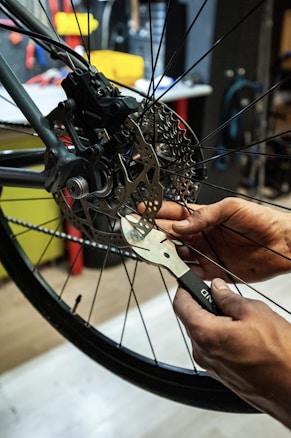 Hands are adjusting a bicycle disc brake with a tool, focusing on the intricate components of the brake system. The background includes a cluttered workshop environment with various tools and equipment visible.