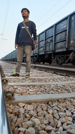 A person stands on a railway track with cargo train cars in the background, wearing a black sweater, jeans, and sneakers with bright green accents. The tracks are covered with gravel and extend into the distance.