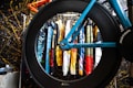 A blue bicycle frame with a large black wheel is displayed, featuring the name 'Airpusher' on the frame and 'Rotor' on the gear. Behind the bike, various surfboards or art pieces with colorful designs are arranged vertically against a backdrop with a chaotic, splattered pattern in black, yellow, and red.