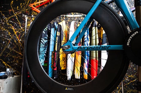 A blue bicycle frame with a large black wheel is displayed, featuring the name 'Airpusher' on the frame and 'Rotor' on the gear. Behind the bike, various surfboards or art pieces with colorful designs are arranged vertically against a backdrop with a chaotic, splattered pattern in black, yellow, and red.