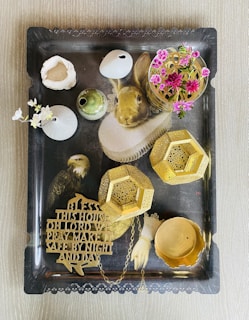 A decorative tray features a well-dressed rabbit painted with a ruffled collar on its surface. Several small objects are placed on the tray, including pink flowers in a vase, gold geometric ornaments, a carved message plaque, and various decorative items resembling vases and bowls. An eagle is also painted on the tray beside the rabbit.