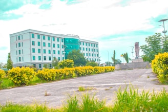 A modern white building with several floors and numerous windows stands prominently. In front of the building, there is a well-maintained garden with vibrant yellow-green bushes lining a paved pathway. A statue on a stone pedestal is located to the right, surrounded by trees and bright sky above.