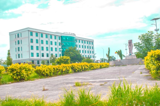 A modern white building with several floors and numerous windows stands prominently. In front of the building, there is a well-maintained garden with vibrant yellow-green bushes lining a paved pathway. A statue on a stone pedestal is located to the right, surrounded by trees and bright sky above.