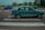 A green car is parked on a pedestrian crossing in an urban area. The scene features a mix of vehicles on the road and buildings in the background, with some trees and a billboard visible. The sky appears overcast, giving the scene a subdued lighting.