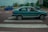 A green car is parked on a pedestrian crossing in an urban area. The scene features a mix of vehicles on the road and buildings in the background, with some trees and a billboard visible. The sky appears overcast, giving the scene a subdued lighting.