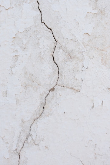 A cracked and weathered wall surface with visible patterns of peeling paint and a prominent vertical fracture running through the center. The wall has a textured appearance with shades of white and light gray.