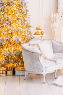 A beautifully decorated Christmas tree adorned with golden ornaments and white lights stands next to a plush, silver-colored sofa. Wrapped gifts in metallic and neutral shades are placed under the tree, enhancing the festive atmosphere. The room features elegant white decor with a hint of classic opulence, including a mantle with candles and gold accents.