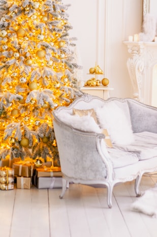 A beautifully decorated Christmas tree adorned with golden ornaments and white lights stands next to a plush, silver-colored sofa. Wrapped gifts in metallic and neutral shades are placed under the tree, enhancing the festive atmosphere. The room features elegant white decor with a hint of classic opulence, including a mantle with candles and gold accents.