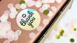 A close-up of a gift box wrapped in brown paper decorated with pink and green circular confetti. A round sticker with 'thank you' written in a cursive font, with a heart above the text, is attached to the paper.