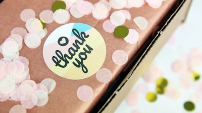A close-up of a gift box wrapped in brown paper decorated with pink and green circular confetti. A round sticker with 'thank you' written in a cursive font, with a heart above the text, is attached to the paper.