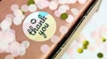 A close-up of a gift box wrapped in brown paper decorated with pink and green circular confetti. A round sticker with 'thank you' written in a cursive font, with a heart above the text, is attached to the paper.