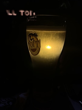 A tall glass filled with beer, illuminated from behind, creating a warm glow against a dark background. The glass has a logo that reads 'TOIT Beer Co.' etched on it.