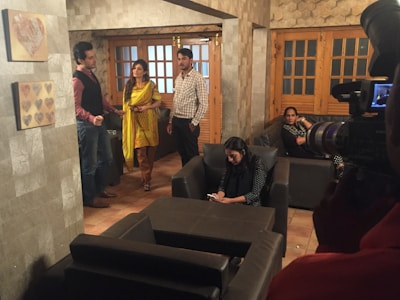 A group of people are in a living room setting with modern decor. The room features stone-textured walls, a heart-themed wall art, several couches, and a large camera setup. Some individuals are standing, while others are seated with one person engaged with a smartphone.