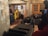 A group of people are in a living room setting with modern decor. The room features stone-textured walls, a heart-themed wall art, several couches, and a large camera setup. Some individuals are standing, while others are seated with one person engaged with a smartphone.