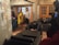 A group of people are in a living room setting with modern decor. The room features stone-textured walls, a heart-themed wall art, several couches, and a large camera setup. Some individuals are standing, while others are seated with one person engaged with a smartphone.