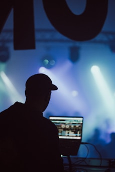 A silhouette of a person wearing a cap is operating a laptop with a DJ software interface. The background is filled with colorful stage lights and smoke, creating an energetic atmosphere.