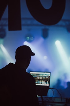 A silhouette of a person wearing a cap is operating a laptop with a DJ software interface. The background is filled with colorful stage lights and smoke, creating an energetic atmosphere.