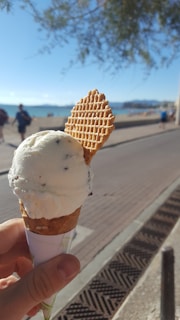 A hand holding a cone of vanilla ice cream topped with a thin waffle biscuit. The background shows a seaside walkway, with the beach and ocean visible. There are people walking along the path, and trees providing shade at the top of the frame.