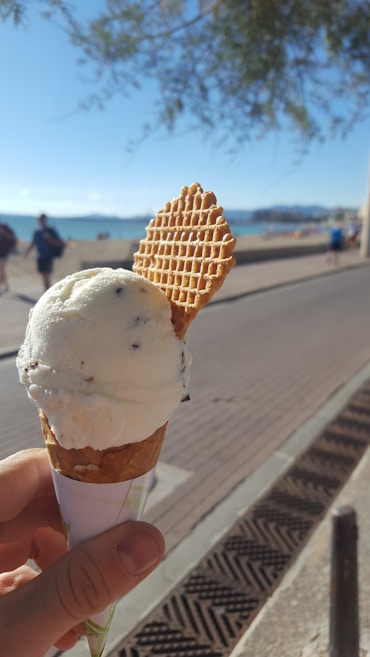 A hand holding a cone of vanilla ice cream topped with a thin waffle biscuit. The background shows a seaside walkway, with the beach and ocean visible. There are people walking along the path, and trees providing shade at the top of the frame.