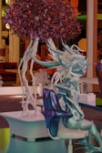 A fantastical, abstract figure resembling a humanoid form with smooth, flowing, and spiky surfaces in various shades of blue, purple, and teal. It appears to be sitting in a modern, stylized setting. A vibrant tree with deep red and purple foliage and white, twisting branches stands beside it. The background features architectural elements and structures, hinting at an imaginative environment.
