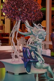 A fantastical, abstract figure resembling a humanoid form with smooth, flowing, and spiky surfaces in various shades of blue, purple, and teal. It appears to be sitting in a modern, stylized setting. A vibrant tree with deep red and purple foliage and white, twisting branches stands beside it. The background features architectural elements and structures, hinting at an imaginative environment.