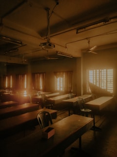 A dimly lit classroom with rows of empty desks and chairs, illuminated by warm sunlight streaming through the windows. The room has a slightly old-fashioned look with ceiling fans and a projector. Curtains are partially drawn, adding to the subdued ambiance.