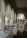 Elegant interior space with long draped curtains lining the windows, allowing soft natural light to filter through. The room features classic wooden furniture, upholstered chairs, and small tables. A patterned carpet decorates the floor, contributing to a luxurious and refined ambiance. Houseplants are placed strategically around the space.