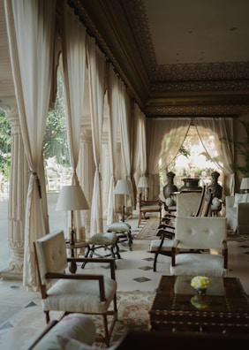 Elegant interior space with long draped curtains lining the windows, allowing soft natural light to filter through. The room features classic wooden furniture, upholstered chairs, and small tables. A patterned carpet decorates the floor, contributing to a luxurious and refined ambiance. Houseplants are placed strategically around the space.