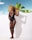 A person wearing a black swimsuit stands confidently against a white wall, with palm trees in the background. They hold a large, colorful sun hat that features a vibrant geometric pattern in shades of orange, yellow, and blue. The sky is clear with a few clouds, creating a tropical atmosphere.