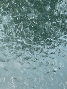 A close-up view of a layer of clear ice cubes with visible texture and light reflections, creating a cool and refreshing appearance.