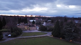 A large estate with multiple historic buildings is surrounded by dense trees. The sky is overcast, with hints of a sunset or sunrise in the distance. The landscape includes a well-maintained lawn and paved pathways. The scene has an atmospheric and serene quality, with minimal artificial lighting visible from the buildings and along the pathways.