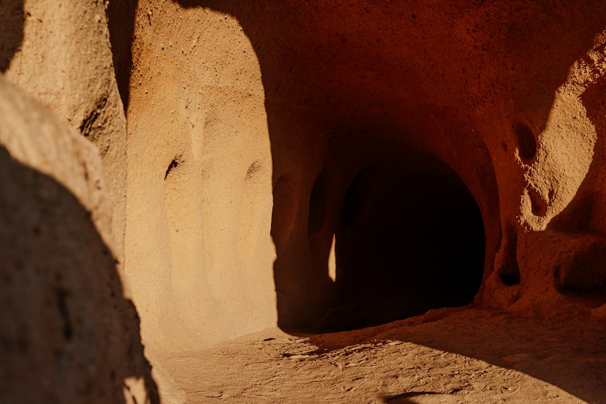 The interior of a space carved into soft rock in sand-orange tones: a rounded, smoothed wall lit warmly from the left, and beside it a dark arched opening leading into an inner tunnel. Recesses and rounded niches are visible in the ceiling and walls, and the floor is covered in dust and crumbling stone