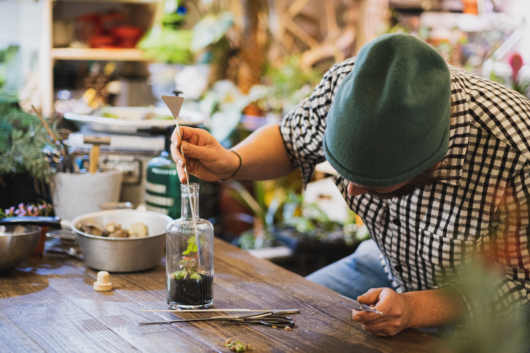 A person in a green beanie lowering a long tool into a narrow glass bottle on a wooden workbench