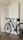 A mountain bike with teal and black accents is positioned indoors, leaning against a white door with geometric designs. The bike has large, rugged wheels suitable for off-road cycling, and a small saddlebag is attached under the seat. The room features a wooden floor and neutral-colored walls, creating a minimalist and clean environment.