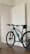 A mountain bike with teal and black accents is positioned indoors, leaning against a white door with geometric designs. The bike has large, rugged wheels suitable for off-road cycling, and a small saddlebag is attached under the seat. The room features a wooden floor and neutral-colored walls, creating a minimalist and clean environment.