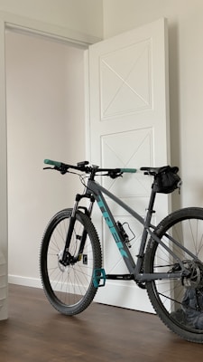A mountain bike with teal and black accents is positioned indoors, leaning against a white door with geometric designs. The bike has large, rugged wheels suitable for off-road cycling, and a small saddlebag is attached under the seat. The room features a wooden floor and neutral-colored walls, creating a minimalist and clean environment.