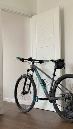 A mountain bike with teal and black accents is positioned indoors, leaning against a white door with geometric designs. The bike has large, rugged wheels suitable for off-road cycling, and a small saddlebag is attached under the seat. The room features a wooden floor and neutral-colored walls, creating a minimalist and clean environment.