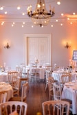 A beautifully decorated room set up for an elegant event, featuring multiple round tables draped with white tablecloths and surrounded by light wooden chairs. Each table is adorned with floral centerpieces and sparkling glassware. A large, ornate chandelier hangs from the ceiling, and warm string lights are draped across the room creating a cozy and inviting atmosphere. Wall-mounted candle sconces enhance the ambiance with soft lighting.