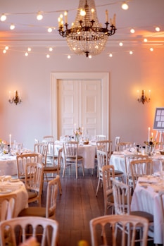 A beautifully decorated room set up for an elegant event, featuring multiple round tables draped with white tablecloths and surrounded by light wooden chairs. Each table is adorned with floral centerpieces and sparkling glassware. A large, ornate chandelier hangs from the ceiling, and warm string lights are draped across the room creating a cozy and inviting atmosphere. Wall-mounted candle sconces enhance the ambiance with soft lighting.