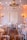 A beautifully decorated room set up for an elegant event, featuring multiple round tables draped with white tablecloths and surrounded by light wooden chairs. Each table is adorned with floral centerpieces and sparkling glassware. A large, ornate chandelier hangs from the ceiling, and warm string lights are draped across the room creating a cozy and inviting atmosphere. Wall-mounted candle sconces enhance the ambiance with soft lighting.