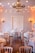 A beautifully decorated room set up for an elegant event, featuring multiple round tables draped with white tablecloths and surrounded by light wooden chairs. Each table is adorned with floral centerpieces and sparkling glassware. A large, ornate chandelier hangs from the ceiling, and warm string lights are draped across the room creating a cozy and inviting atmosphere. Wall-mounted candle sconces enhance the ambiance with soft lighting.