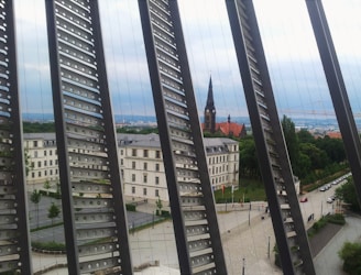 Metal beams with circular cutouts create a pattern in the foreground, framing a view of a historical building with an ornate spire and red-tiled roof. The scene includes leafy trees and cloudy skies, suggesting an urban environment with a mix of architecture and greenery.